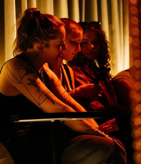 Three people sitting closely together in warm, low lighting, one with visible tattoos on their arms.