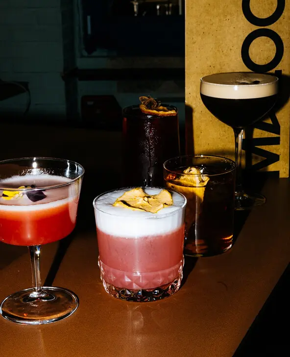 Five assorted cocktails on a brown surface, including two with foam and dried garnishes and one dark coffee-based drink in a coupe glass.