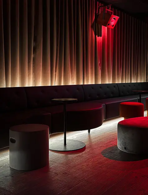 Dimly lit lounge with black tufted seating, round tables and stools, illuminated by soft backlighting and red accent lights.