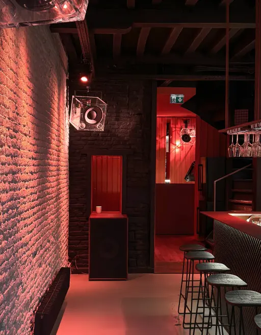 Interior of a dimly lit bar with red lighting, black stools lined up by a counter, exposed brick walls, and hanging glassware above the bar.