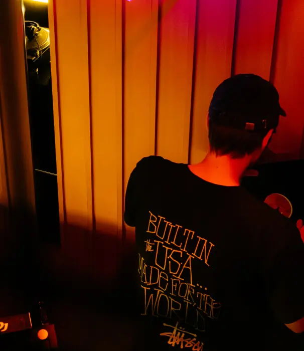 Person wearing a black cap and black shirt with text reading 'BUILT IN the USA... MADE FOR THE WORLD' under warm red and yellow lighting.