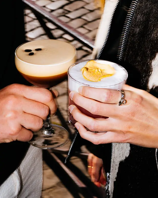 Two hands holding different cocktails, one with frothy espresso and three coffee beans on top, the other with foam and a dried citrus garnish.