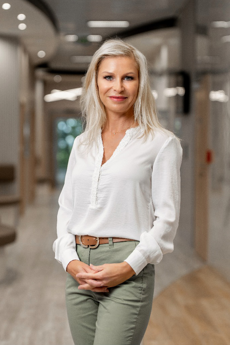 Žena s blond vlasy a bílou blůzou stojící v moderní kanceláři s rukama sepjatýma před tělem.