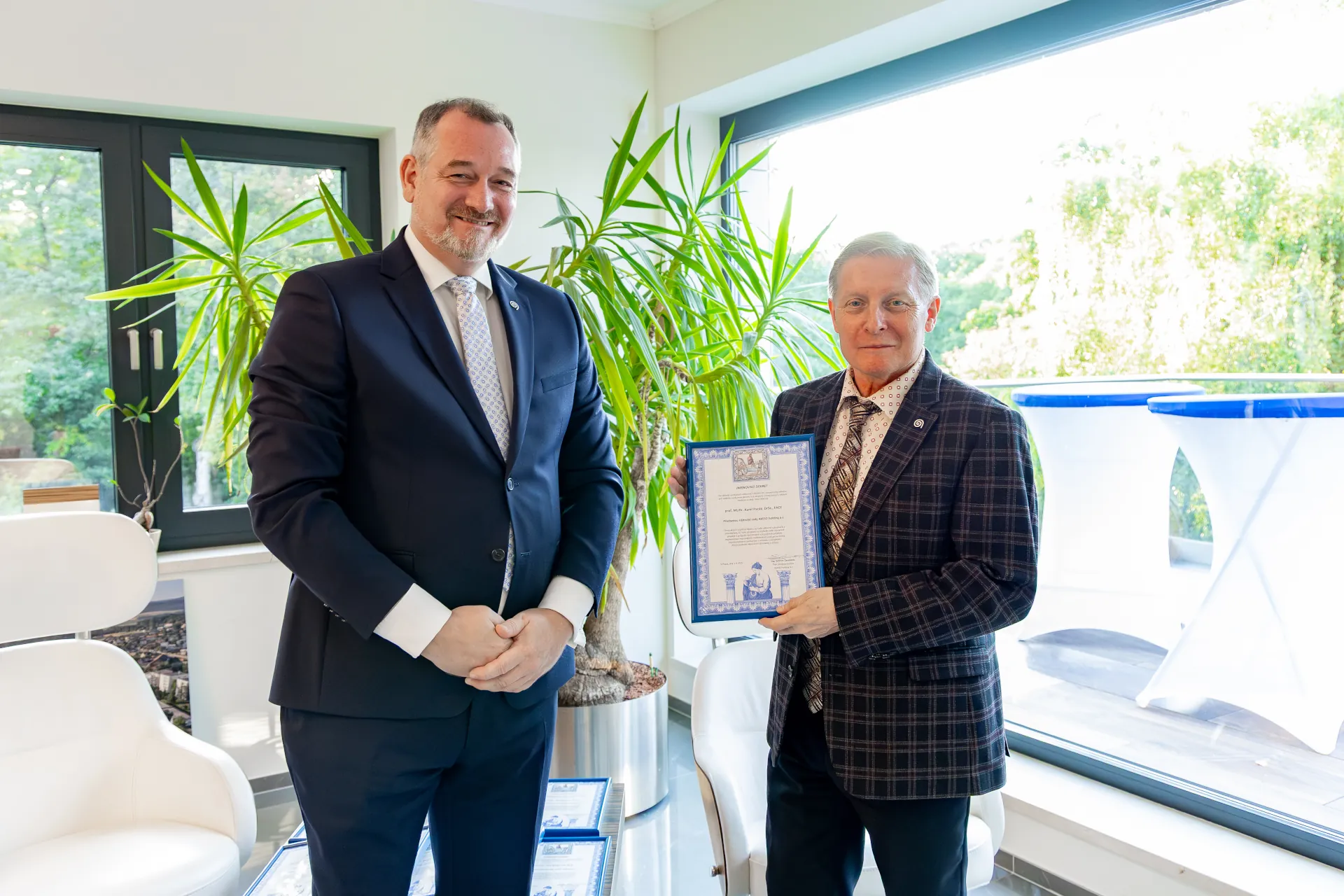 Dva muži ve formálním oblečení stojí v místnosti, jeden drží certifikát, v pozadí je zelená rostlina a velké okno s výhledem na venkovní terasu.