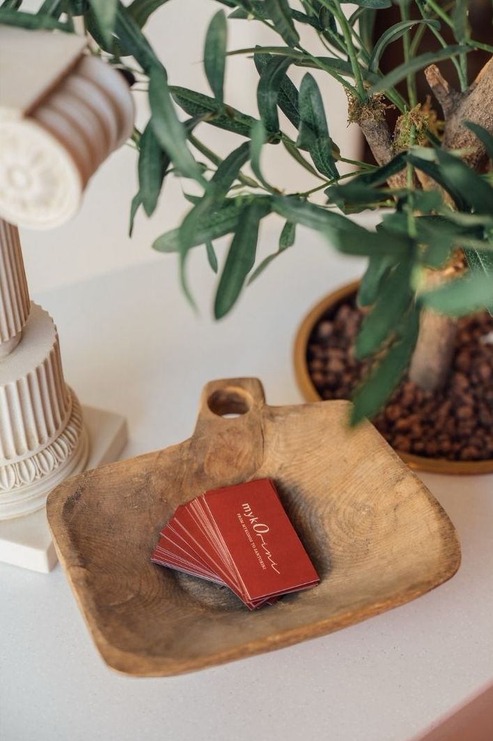 A wooden dish holding red business cards sits on a white surface next to a decorative plant and a column-shaped object.