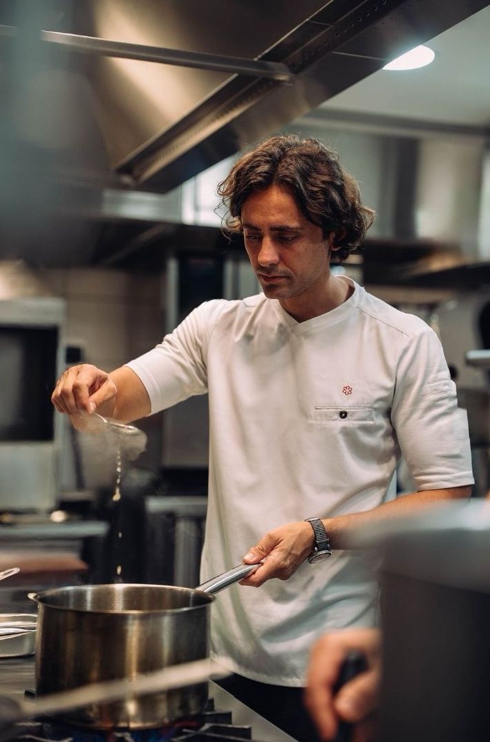 A chef in a white shirt adding ingredients to a pot in a professional kitchen. The kitchen has stainless steel appliances and a well-organized workspace.