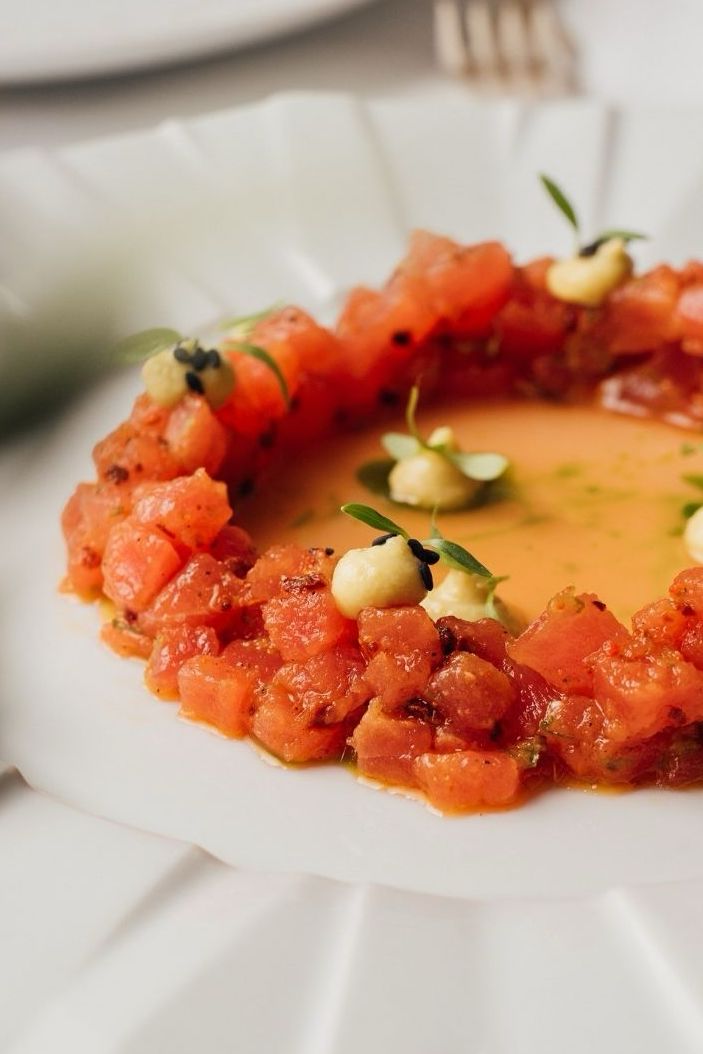 A gourmet dish featuring diced salmon arranged in a circular pattern on a white plate. The salmon is surrounded by a light sauce and garnished with small dollops of cream and green sprouts.