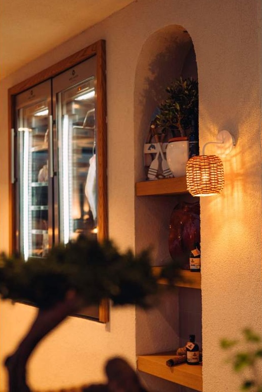 A cozy interior view of a wall with built-in shelves displaying decorative items, including a potted plant. A woven light fixture casts warm light, with a glass door refrigerator visible in the background.