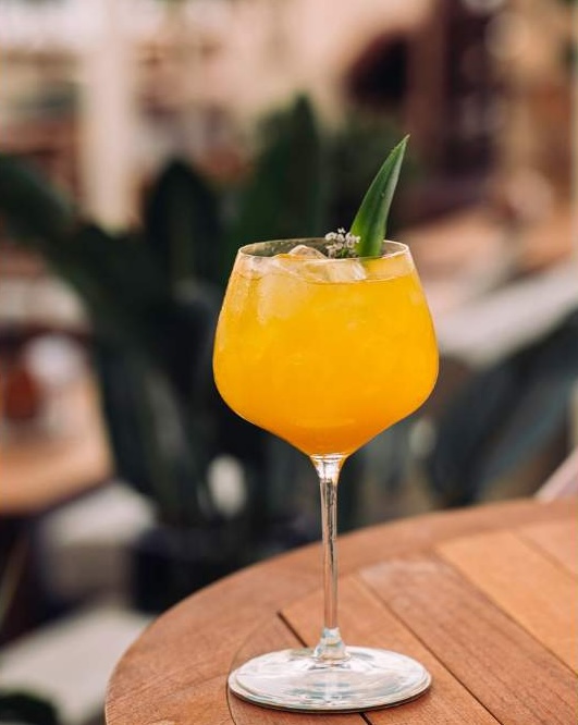 A bright orange cocktail in a large stemmed glass, garnished with a green leaf. The drink contains ice and is placed on a wooden table with soft focus background elements, including greenery.