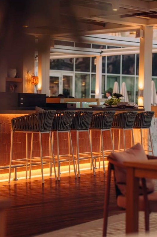 A view of a modern bar area with high stools, warm lighting, and wooden flooring. The background features large windows, providing a glimpse of an outdoor area with umbrellas.