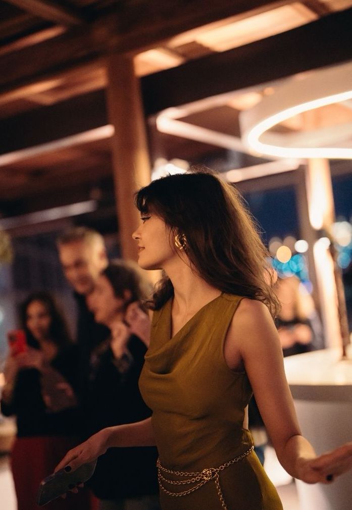 A woman with long hair wearing a green dress and gold accessories stands in a elegantly lit venue. Behind her, a group of people are engaged in conversation, with some holding smartphones.