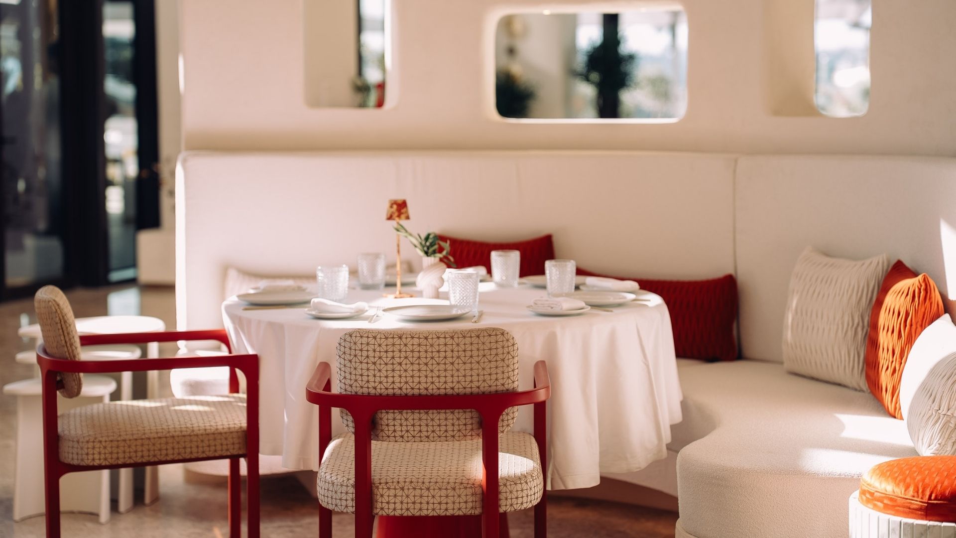 A cozy restaurant setting with a round table set for dining, surrounded by upholstered chairs. Sunlight enters through large windows, creating a bright atmosphere.