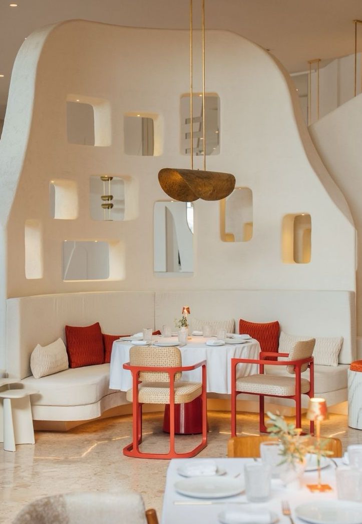 An elegant dining area in a modern restaurant featuring a white wall with cut-out shelves. A round table is set for two, surrounded by cream-colored and red chairs, with soft lighting and decorative elements.