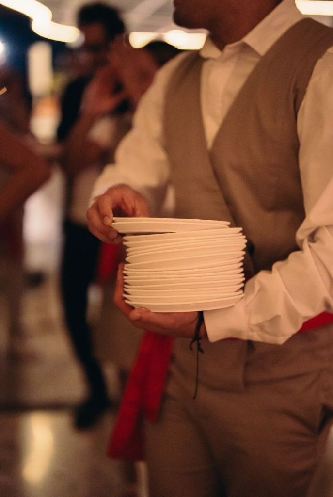 A person in a light-colored vest holding a stack of white plates at a gathering, with blurred figures and soft lighting in the background.