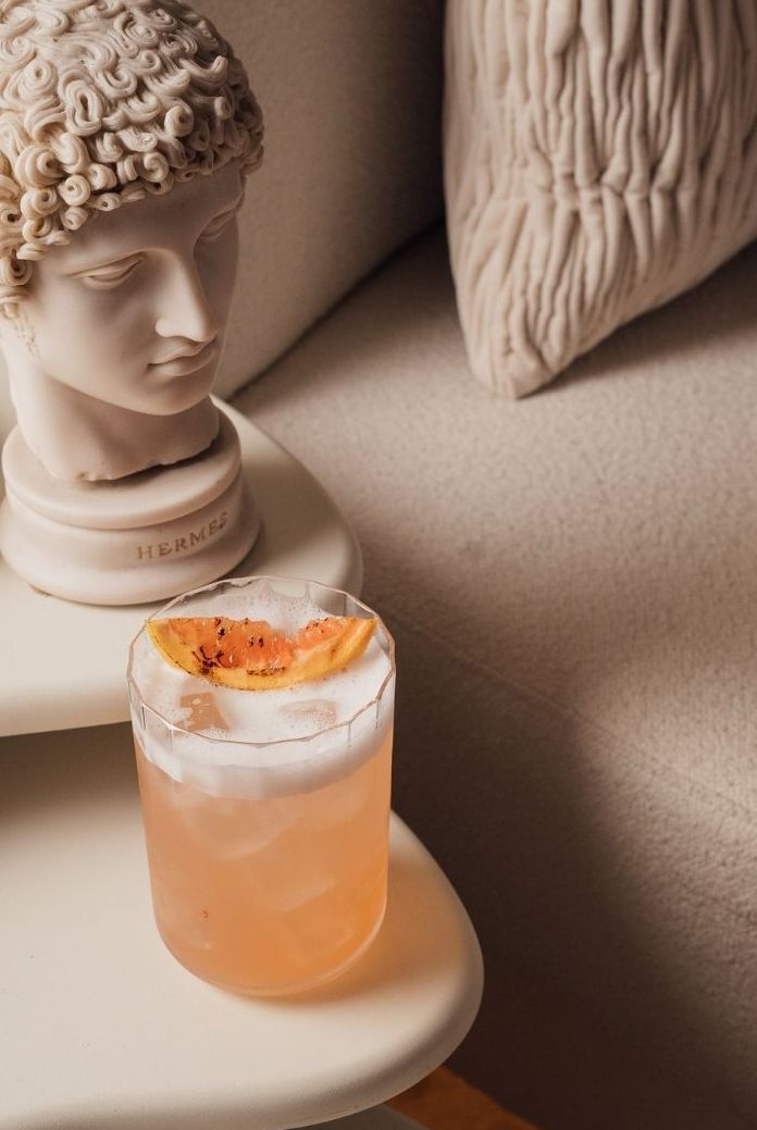A decorative drink with a peach slice on top, placed on a side table next to a white bust sculpture. The background features a textured cream-colored fabric.