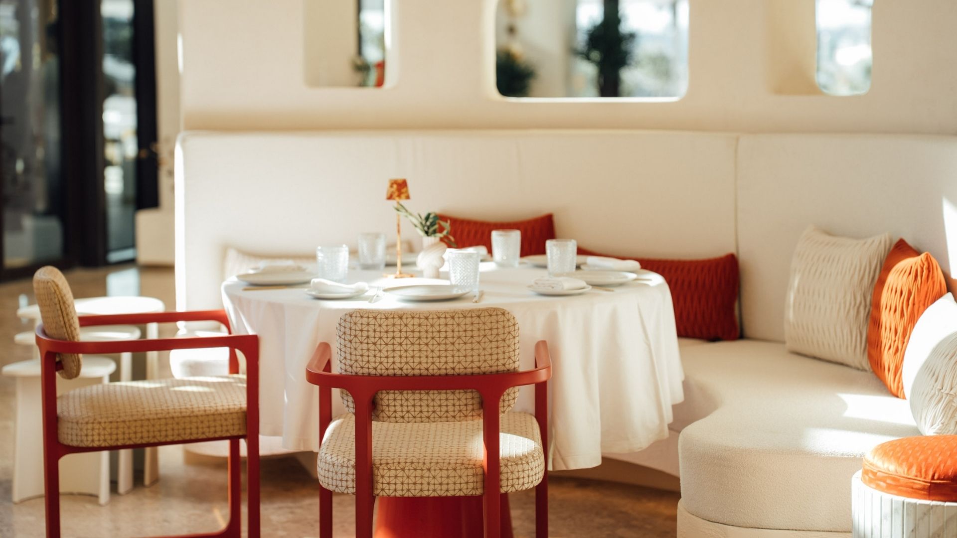 A cozy dining area featuring a round table set for a meal, surrounded by woven chairs. The decor includes a white couch with orange cushions and subtly lit by natural light.