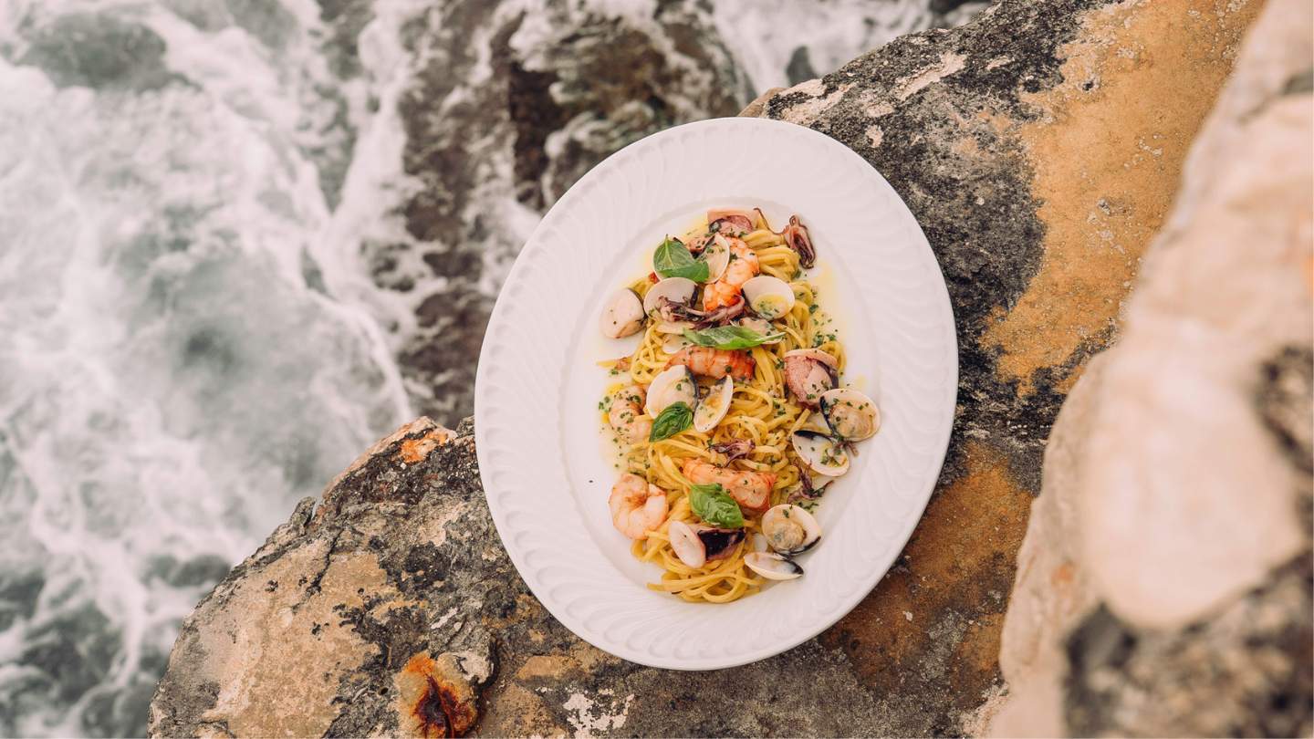 Seafood pasta dish with shrimp, clams, and basil on rocky beach by ocean waves