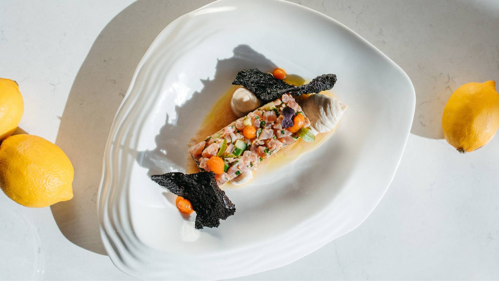 Fine dining tuna tartare with diced vegetables, seaweed garnish, pearl, and lemon on white plate