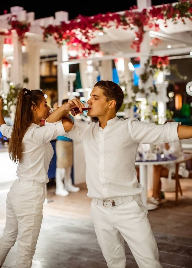 Couple in white attire dancing at festive outdoor event with red flower decorations overhead