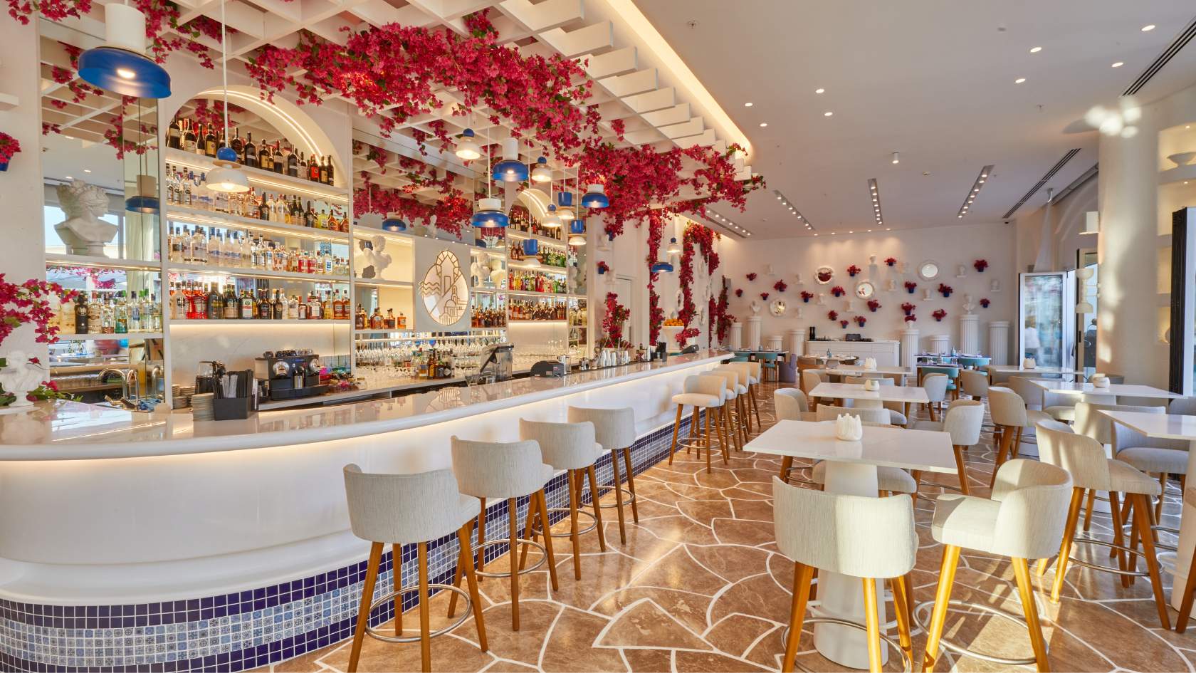 Modern restaurant interior with cream bar stools, white tables, red floral ceiling decorations, and well-stocked bar shelving.