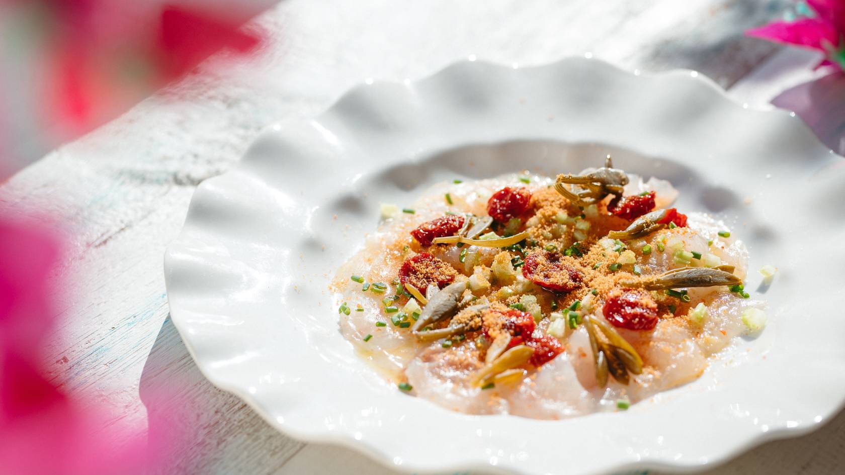Elegantly plated fish dish with red berries, capers, and herbs on white scalloped plate.