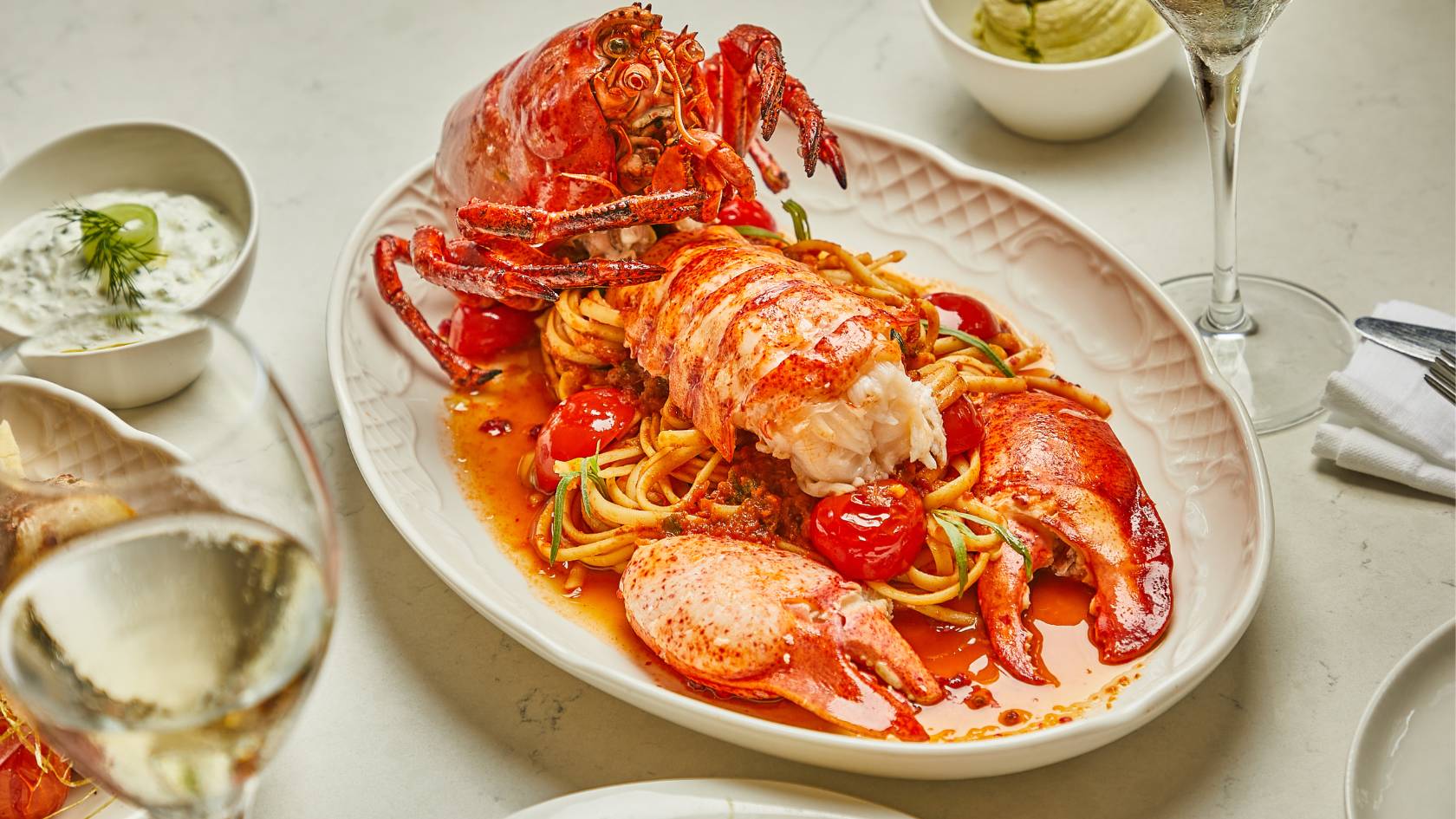 Cooked lobster on white plate with pasta, sauces, and wine glass
