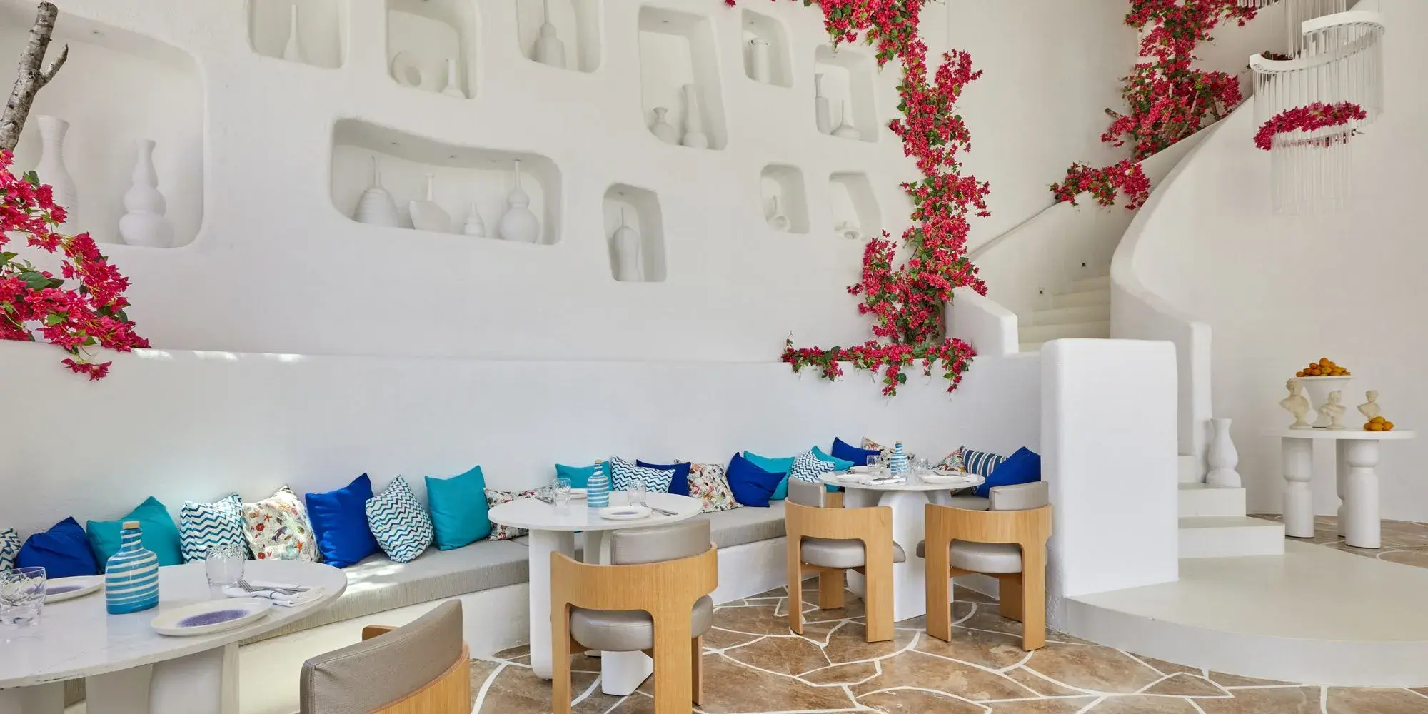 White Mediterranean restaurant with bright magenta bougainvillea flowers, colorful blue cushions, and wooden chairs.