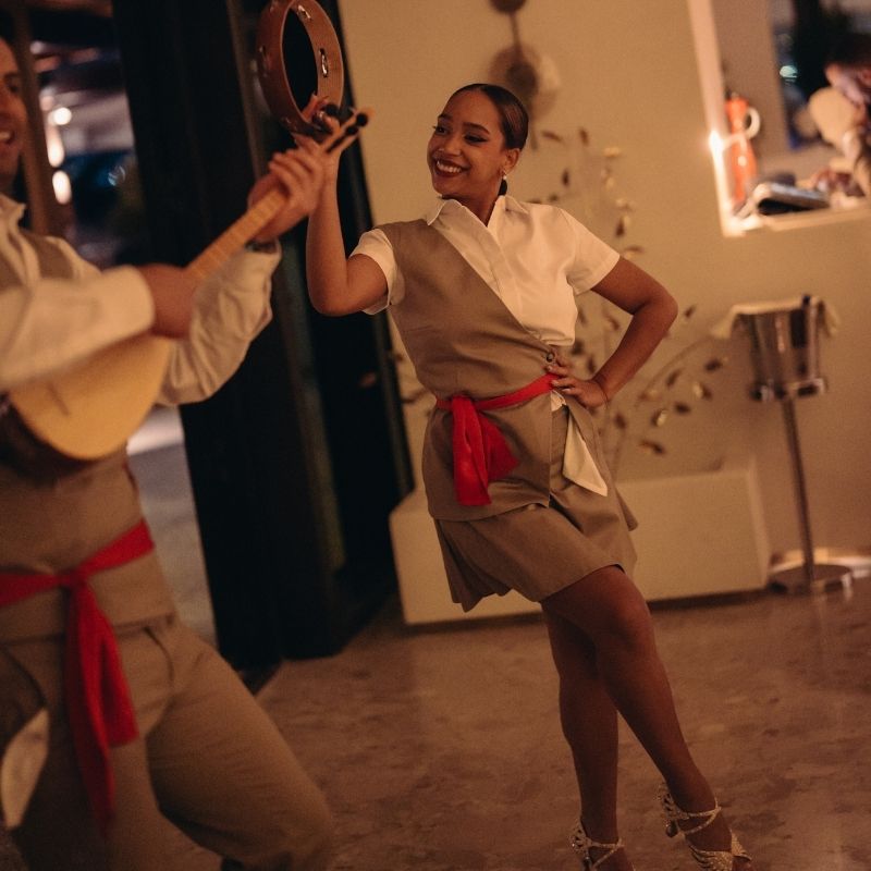 Smiling woman in beige dress with red sash dancing with tambourine at party.