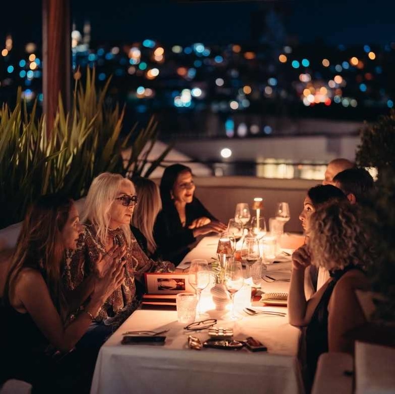 Group of women dining outdoors at night with city lights and candles illuminating the table.