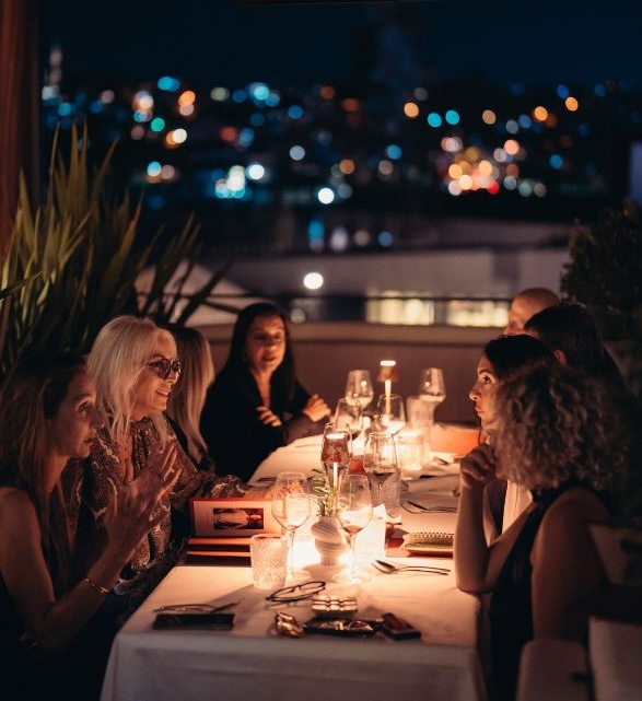 Women dining outdoors at night with city lights and plants in background.