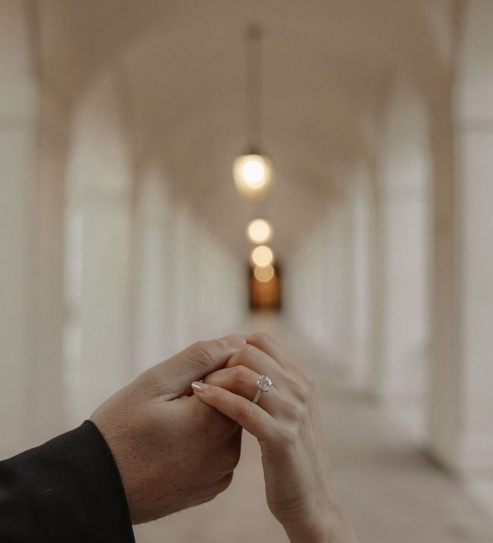 Couple's hands together displaying diamond engagement ring in elegant hallway with soft lighting