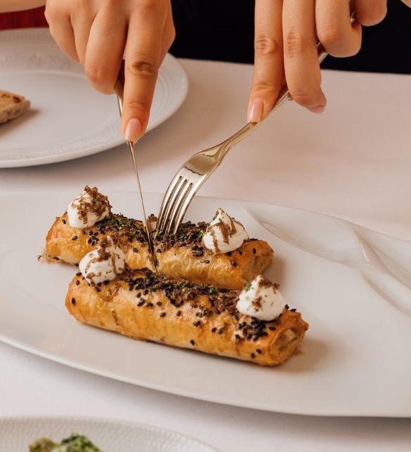 Hands cutting into elegant pastry dessert with cream and black sesame seeds on plate