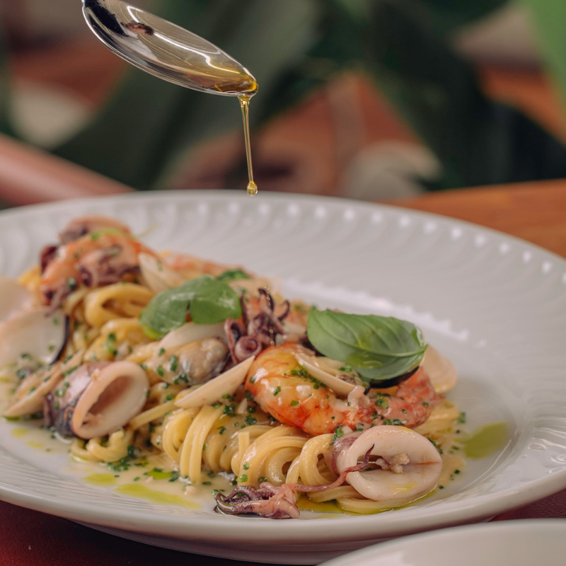 Olive oil drips onto plated pasta with squid, shrimp, and fresh basil garnish