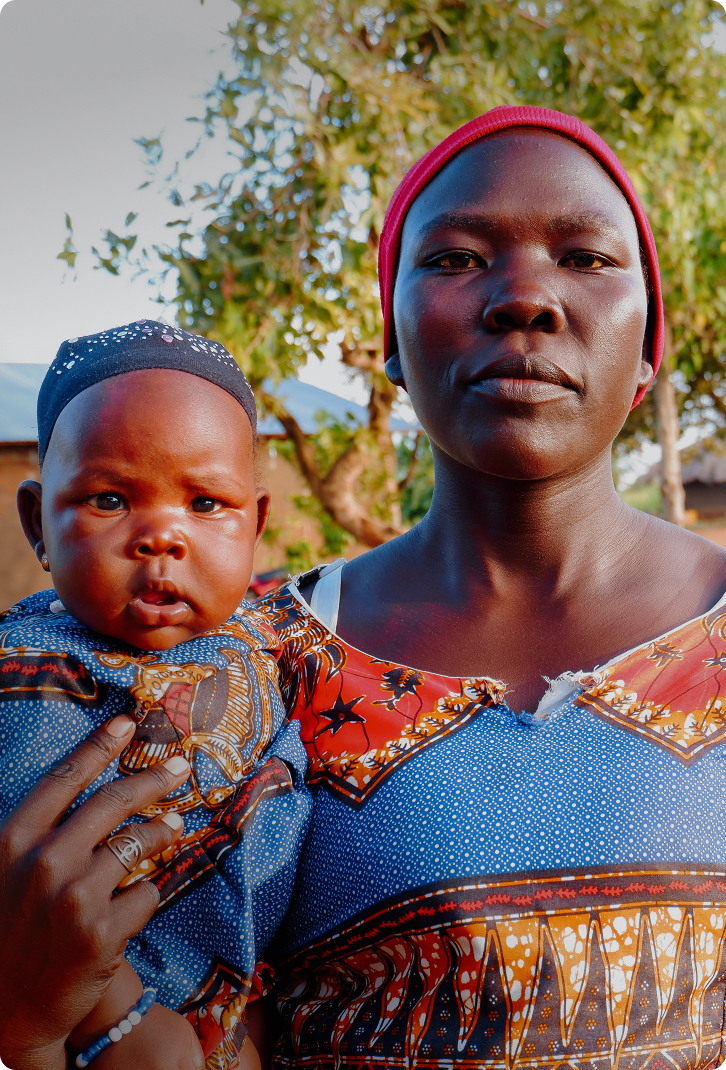 Portrait of a woman holding a baby, both wearing colorful patterned clothing, standing outdoors in front of trees and traditional houses in Uganda, Africa.