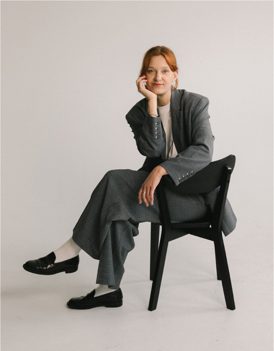 A woman with red hair sits relaxed on a chair, wearing a gray suit with a white shirt and black loafers, resting her head on her hand.