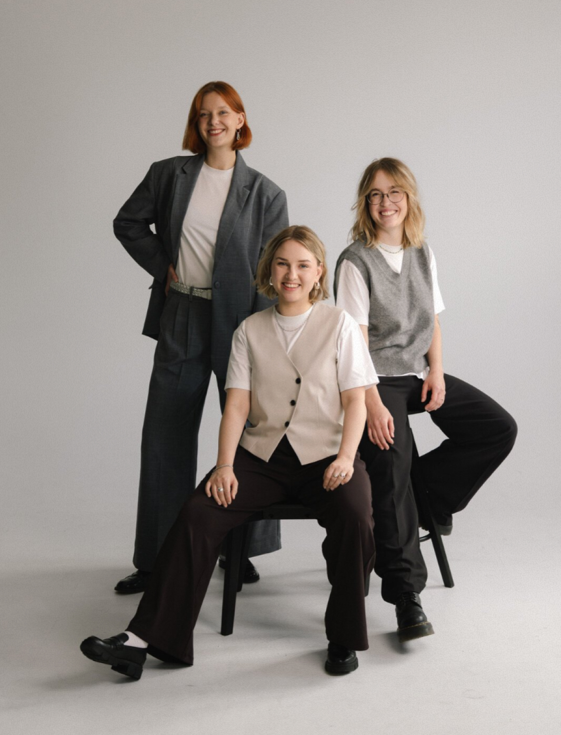 Three women pose together in the studio. One stands in a gray suit, while the other two sit on chairs wearing light vests over white T-shirts with dark trousers.