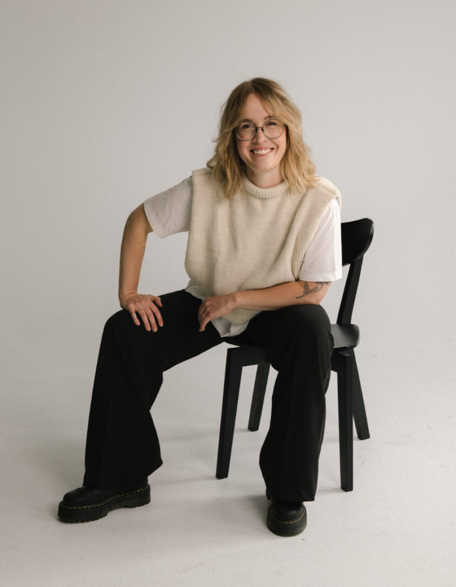 A woman with blonde hair and glasses sits casually on a chair, wearing a white T-shirt with a knitted vest, black trousers, and platform shoes, smiling at the camera.