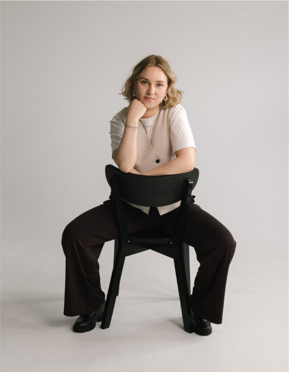 A woman with blonde hair sits backwards on a chair, wearing a beige vest over a white T-shirt with dark trousers and black shoes.