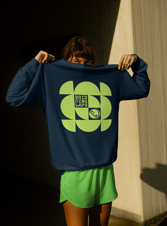 Person, die ein marineblaues Bridging Gaps-Sweatshirt mit einem hellgrünen geometrischen Logo-Design auf der Vorderseite hochhält, das bei natürlichem Licht dargestellt wird.