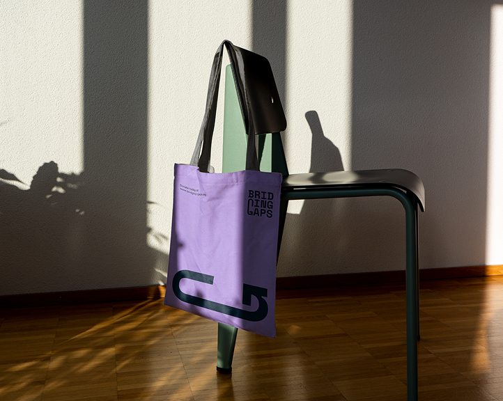 Eine lilafarbene Bridging Gaps-Tragetasche mit dem Logo und dem Spendenzyklussymbol, die im natürlichen Sonnenlicht an einem Stuhl hängt.