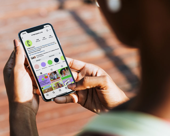 Nahaufnahme von Händen, die ein Smartphone halten, auf dem das Instagram-Profil von Bridging Gaps zu sehen ist, auf dem die visuelle Identität der Marke und farbenfrohe Beiträge zu sehen sind.