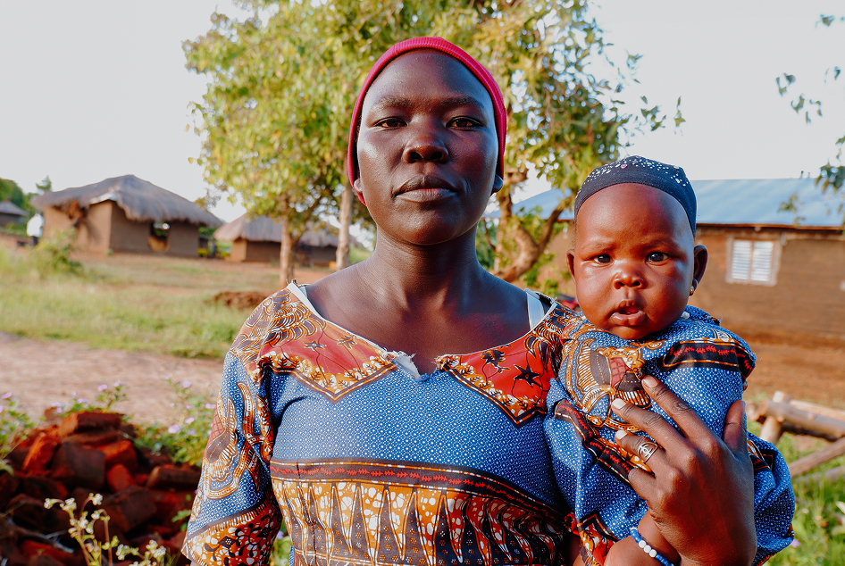 Porträt einer Frau mit einem Baby, beide in bunt gemusterter Kleidung, die draußen vor Bäumen und traditionellen Häusern in Uganda, Afrika, steht.