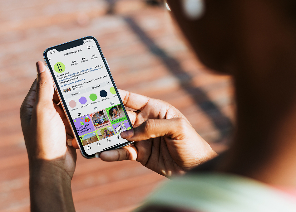 Nahaufnahme von Händen, die ein Smartphone halten, auf dem das Instagram-Profil von Bridging Gaps zu sehen ist, auf dem die visuelle Identität der Marke und farbenfrohe Beiträge zu sehen sind.