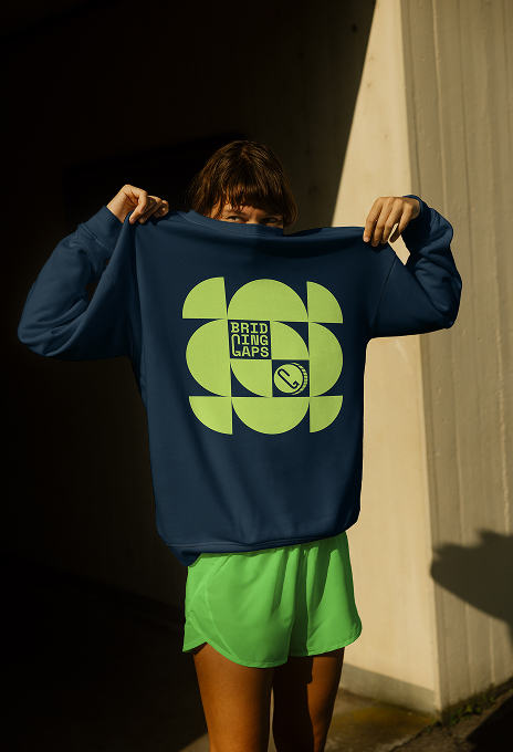Person, die ein marineblaues Bridging Gaps-Sweatshirt mit einem hellgrünen geometrischen Logo-Design auf der Vorderseite hochhält, das bei natürlichem Licht dargestellt wird.