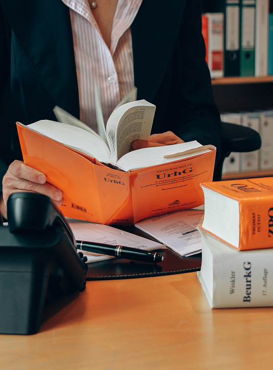 Person an einem Schreibtisch, die ein juristisches Nachschlagewerk mit orangefarbenem Einband liest, umgeben von juristischen Büchern und Büromaterial, das juristische Expertise symbolisiert.