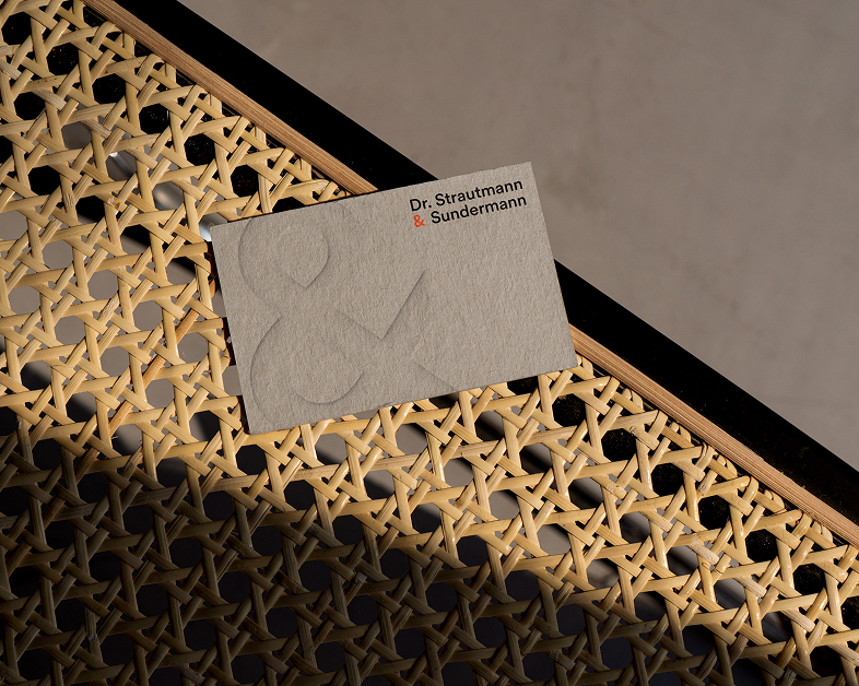 Eine Nahaufnahme der Visitenkarte des Unternehmens auf einem Stuhl aus Zuckerrohr mit dem geprägten Und-Symbol und dem aufgedruckten Logo „Dr. Strautmann & Sundermann“ in Schwarz und Orange.