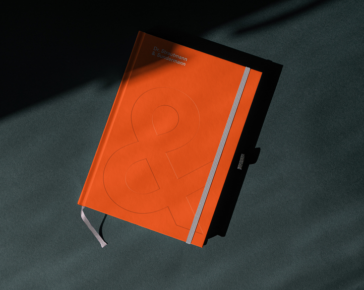 Leuchtend orangefarbenes Notizbuch mit geprägtem Und-Zeichen und dem Dr. Strautmann & Sundermann-Logo, fotografiert in dramatischem Licht und Schatten.
