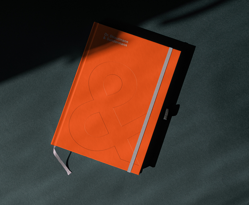 Leuchtend orangefarbenes Notizbuch mit geprägtem Und-Zeichen und dem Dr. Strautmann & Sundermann-Logo, fotografiert in dramatischem Licht und Schatten.