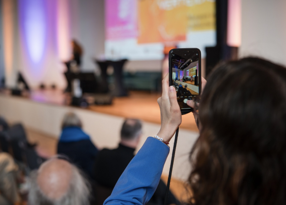 Konferenzszene mit Publikum, Bühne und einem Teilnehmer, der ein Foto auf seinem Handy bei der Veranstaltung Stellung beziehen, Demokratie verteidigen! macht.