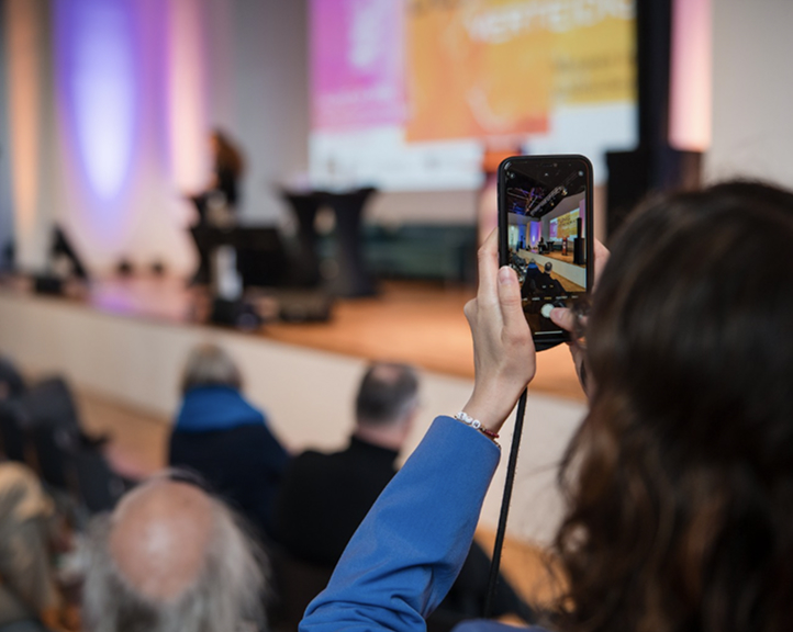 Konferenzszene mit Publikum, Bühne und einem Teilnehmer, der ein Foto auf seinem Handy bei der Veranstaltung Stellung beziehen, Demokratie verteidigen! macht.